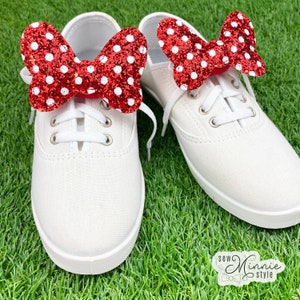 May include: A pair of white canvas shoes with red and white polka dot glitter bows attached to the laces. The shoes are on a green grassy surface.