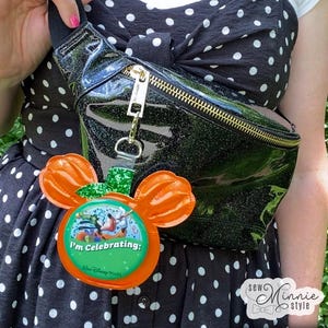 May include: Black glitter fanny pack with a gold zipper and hardware. Attached is a Mickey Mouse-shaped charm with orange ears and the text "I'm Celebrating: Walt Disney World". The bag is worn over a black and white polka dot dress.