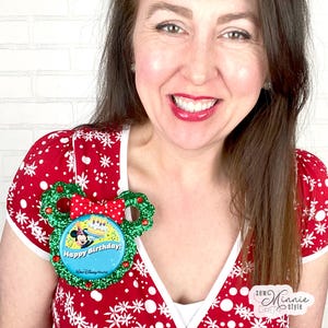 May include: A festive, glittery Mickey Mouse-shaped pin with a red bow and a blue button that says "Happy Birthday!" and "Walt Disney World." The pin is green and red, and the background is a red top with white snowflakes.