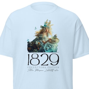 Puede incluir: Camiseta azul claro con un gráfico de una mujer con un sombrero blanco y un vestido azul que sostiene una cesta de pájaros. El texto "1829 Collectors Edition Alice Pleasance Liddell Alice" está impreso debajo del gráfico.