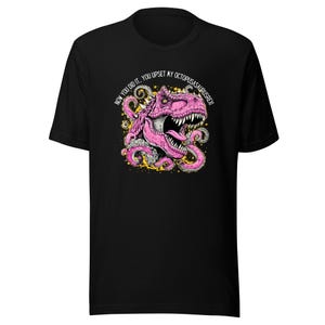 Puede incluir: Camiseta negra con un gráfico híbrido de pulpo-dinosaurio rosa y el texto "NOW YOU DID IT... YOU UPSET MY OCTOPUSASAURUSREX!!". El diseño incluye detalles en amarillo y dorado.