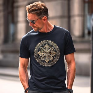May include: A navy blue t-shirt featuring a gold-colored, circular design with intricate patterns. The design is centered on the chest. The shirt has short sleeves and a crew neck.