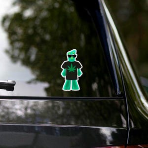 May include: A green sticker of a cartoon figure wearing sunglasses and a black t-shirt with a cannabis leaf design. The figure is standing with its arms at its sides and is wearing a green hat and green pants. The sticker is affixed to a car window.