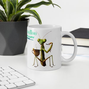 May include: White ceramic mug with a green praying mantis holding a cup of coffee. The mug has the text "Coffee with Mantis" printed on it.