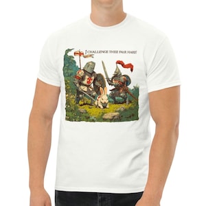 Op de afbeelding: Wit T-shirt met een humoristische illustratie van twee ridders in harnas die een konijn uitdagen. De tekst "I CHALLENGE THEE FAIR HARE!" staat erboven. De ridders zijn gewapend met zwaarden en schilden, en het konijn staat in het midden.
