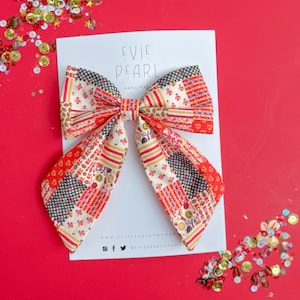 Op de afbeelding: Een handgemaakte Evie Pearl haarstrik met een patchwork design in rood, crème, zwart en geel. De strik ligt op een witte kaart met het Evie Pearl logo en social media handles. Decoratieve pailletten zijn verspreid.