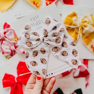 May include: White fabric hair bow with a brown football pattern. The bow is tied in a large, full loop. The bow is displayed on a white card with the text "Evie Pearl Handmade" and social media handles.