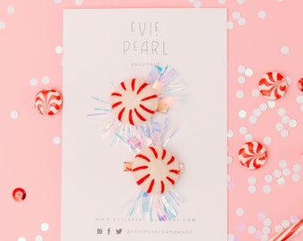 May include: Two candy cane-themed hair clips on a white card with the Evie Pearl Handmade logo, surrounded by peppermint candies and confetti on a pink background. The clips feature a red and white peppermint design with gold-coloured clips.