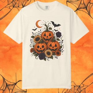 Puede incluir: Una camiseta de manga corta color crema con un gráfico central de tres calabazas de Halloween naranjas rodeadas de girasoles, rosas de color morado oscuro, hojas verdes, una luna creciente y murciélagos volando sobre un patrón de ondas.
