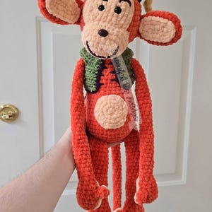 May include: A handmade crochet monkey doll in shades of orange, tan, green, and purple. The monkey has long arms and legs, large ears, and a smiling face. It wears a green vest and a purple hat. The text "Graceful By Ashiya" is visible.