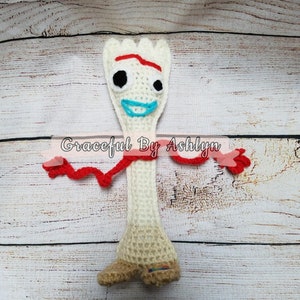 May include: A crocheted toy fork with a red scarf, blue eyes, and a blue mouth. The toy is standing on a wooden surface. The text "Graceful By Ashlyn" is visible in the image.