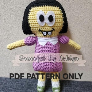 May include: Crocheted doll with a yellow face, black hair, and a purple dress. The doll has white eyes, a smiling mouth, and small glasses. The text "Graceful By Ashlyn" and "PDF PATTERN ONLY" are displayed on the image.