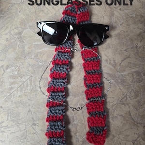 May include: A red, gray, and black crocheted scarf with black sunglasses and a silver chain. The text "SCARF AND SUNGLASSES ONLY" is displayed above the items. The scarf is made of alternating red and gray stripes.