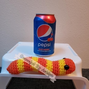 May include: A crocheted yellow and orange worm with black eyes sits on a white surface next to a can of Pepsi Wild Cherry soda.