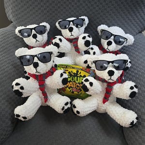 May include: Five handmade white crochet teddy bears wearing black sunglasses and red, gray, and white scarves. The bears have black paws and are holding a bag of Sour Patch Kids candy.
