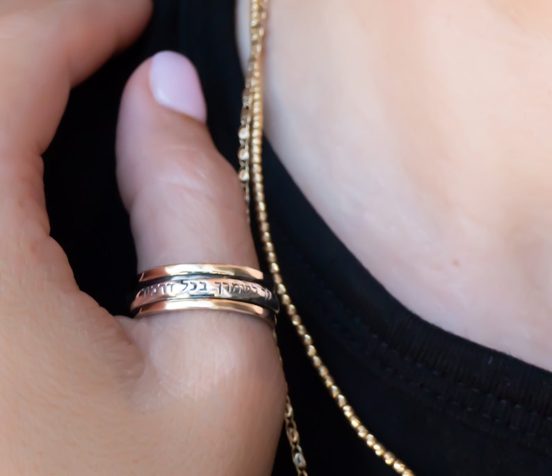 May include: A silver spinning ring with Hebrew text inscribed on the band. The ring is worn on a person's finger.