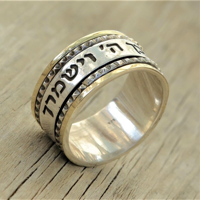 May include: A silver and gold spinning ring with Hebrew text inscribed around the band. The ring has a textured edge.