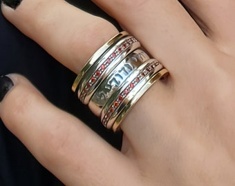 Könnte beinhalten: Ein Silber- und Goldring mit roten Edelsteinen. Der Ring besteht aus mehreren Bändern, die sich umeinander drehen.