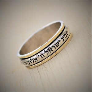 May include: A silver and gold spinning ring with Hebrew text inscribed on the band. The text reads "Shema Yisrael Adonai Eloheinu Adonai Echad" which translates to "Hear O Israel, the Lord our God, the Lord is One."
