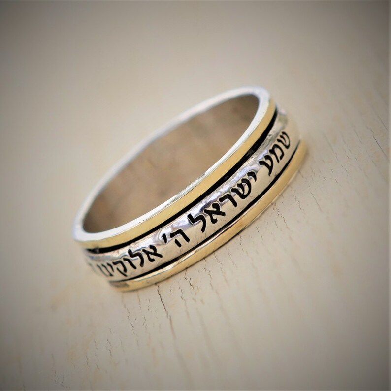 May include: A silver and gold spinning ring with Hebrew text inscribed on the band. The text reads "Shema Yisrael Adonai Eloheinu Adonai Echad" which translates to "Hear O Israel, the Lord our God, the Lord is One."