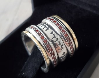 May include: A silver spinning ring with a gold band and red gemstones. The ring has Hebrew writing on it.