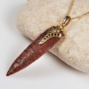 May include: A pendant necklace featuring a reddish-brown arrowhead pendant with a gold-colored metal setting. The setting is adorned with small black gemstones. The necklace chain is also gold-colored.