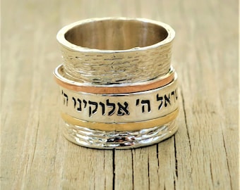 May include: A silver-toned ring with a textured surface and three bands of different colors. The middle band has Hebrew text. The ring is set on a wooden surface.