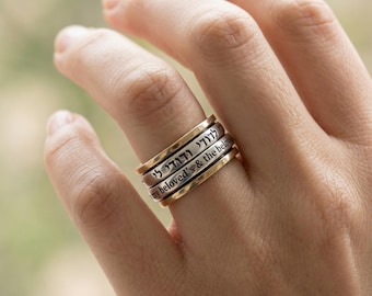 May include: A stack of three rings with gold and silver bands. The rings have engraved text in Hebrew and English. The rings are worn on a finger. The background is blurred.