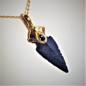 May include: A gold pendant necklace with a black arrowhead charm. The arrowhead is set with a green gemstone and surrounded by small red gemstones.