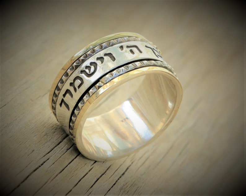 May include: A silver and gold spinning ring with Hebrew text inscribed on the band. The ring has a textured edge.