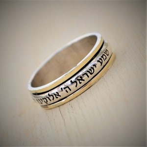 May include: A silver and gold spinning ring with Hebrew text inscribed on the band. The text reads "Shema Yisrael Adonai Eloheinu Adonai Echad" which translates to "Hear O Israel, the Lord our God, the Lord is One."