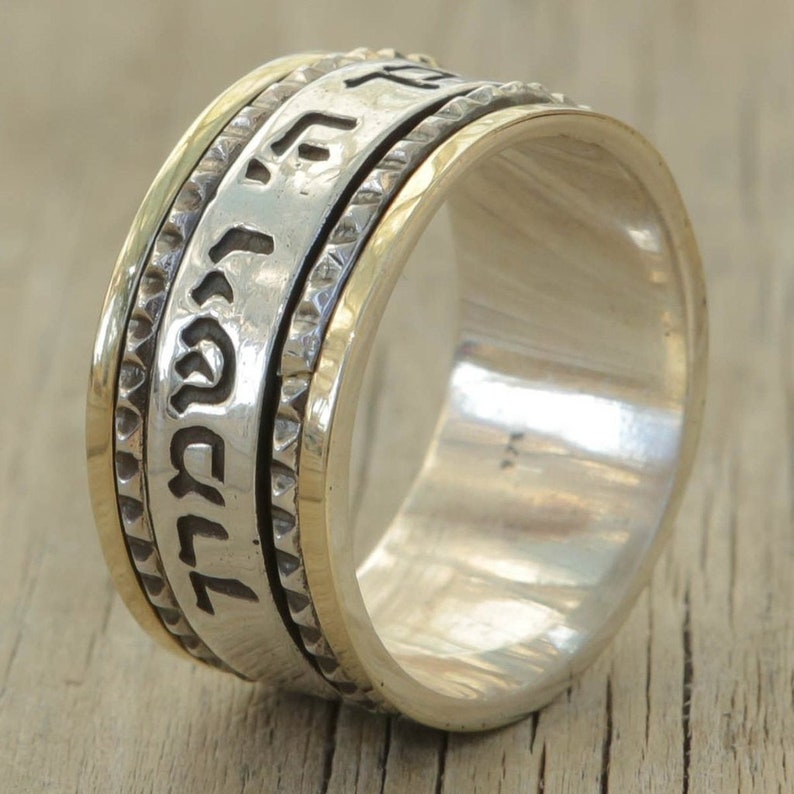 May include: A silver and gold spinning ring with Hebrew text inscribed on the band. The ring is on a wooden surface.