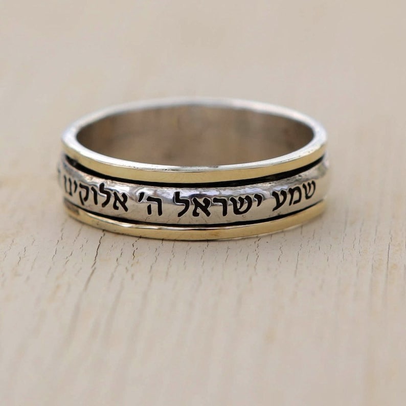 May include: A silver and gold spinning ring with Hebrew text that reads "Shema Yisrael Adonai Eloheinu Adonai Echad" which translates to "Hear O Israel, the Lord our God, the Lord is One".
