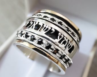 May include: A silver and gold spinning ring with black Hebrew text and black gemstones. The ring has a wide band and is designed to spin.