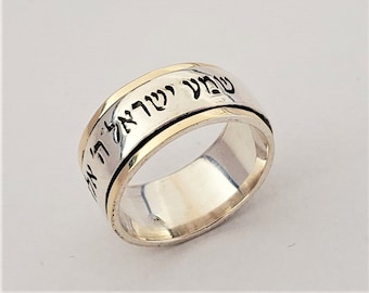 May include: A silver ring with a gold band and Hebrew text. The ring has a spinning outer band. The text is in black and is visible on the gold band. The ring is set against a white background.