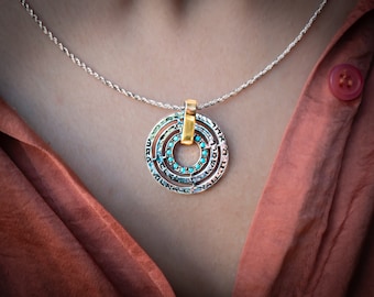 May include: Silver-toned pendant necklace with a gold-colored bail. The pendant features concentric circles with turquoise-colored stones and Hebrew text. The necklace is displayed on a person wearing a rust-colored top.