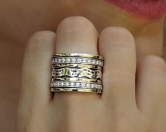 Könnte beinhalten: Ein Silber- und Goldring mit mehreren Bändern, jedes mit einem anderen Design. Das obere Band hat eine Reihe kleiner, klarer Steine und hebräische Schriftzeichen. Der Ring wird an einem Finger getragen.