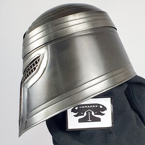 May include: A silver helmet with a mesh face mask and a black fabric background. The helmet has a raised design on the front and a black sticker with the word "CARAPAX" on it.