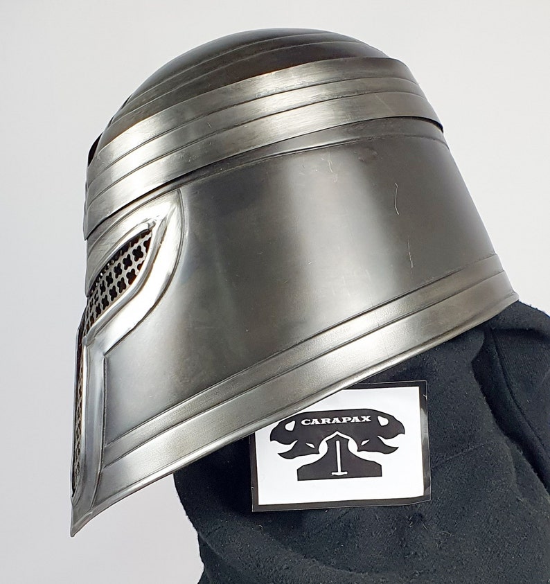 May include: A silver helmet with a mesh face mask and a black fabric background. The helmet has a raised design on the front and a black sticker with the word "CARAPAX" on it.