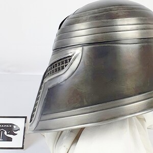 May include: A silver helmet with a black mesh visor and a series of raised ridges on the top. The helmet is made of metal and has a worn, weathered look. The helmet is sitting on a white surface. The text "CARAPAX" is visible on a sticker in the lower left corner of the image.