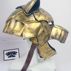 May include: A gold metal helmet with a wolf head design. The helmet has a leather strap for fastening.