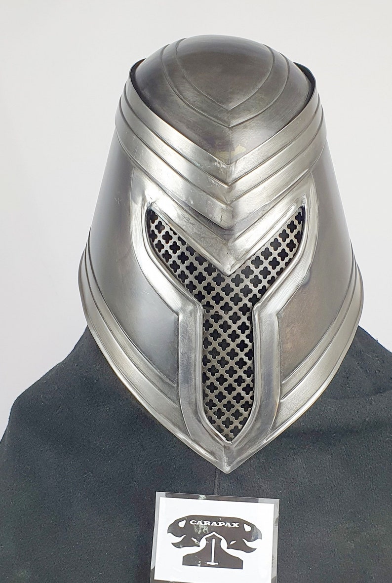 May include: A silver metal helmet with a mesh face mask and a raised design on the top. The helmet is on a black surface with a white label that says "CARAPAX".