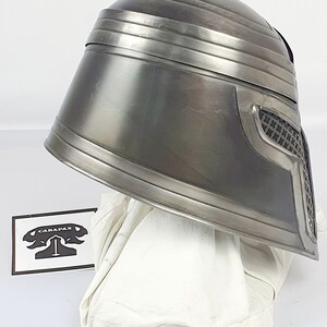 May include: A silver metal helmet with a textured surface and a black mesh visor. The helmet is sitting on a white fabric surface.