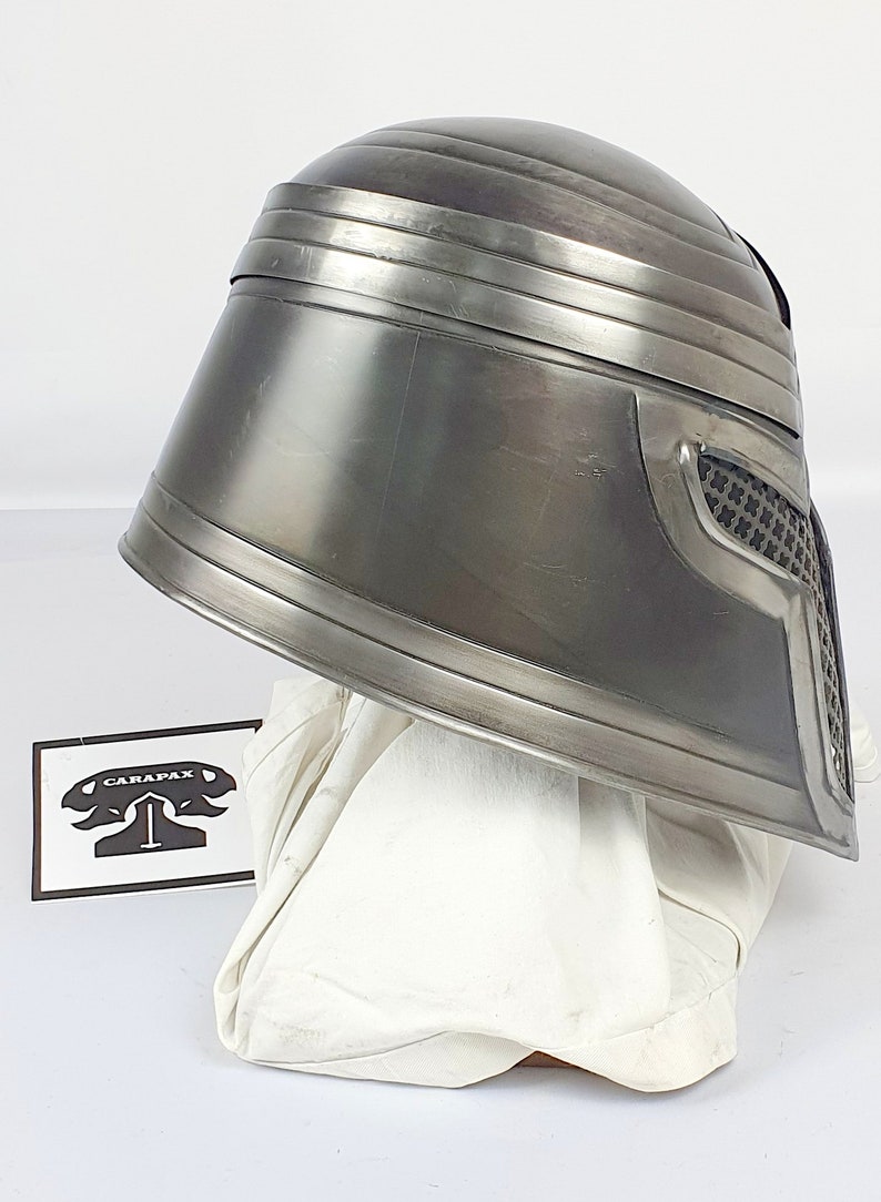 May include: A silver metal helmet with a textured surface and a black mesh visor. The helmet is sitting on a white fabric surface.
