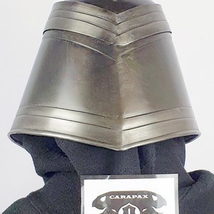 May include: A dark gray metal helmet with a pointed top and a series of ridges around the brim. The helmet is sitting on a black fabric surface. The text "CARAPAX" is visible on a white sticker in the lower right corner of the image.