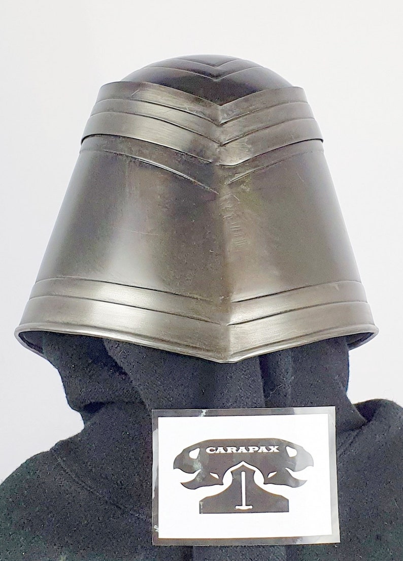 May include: A dark gray metal helmet with a pointed top and a series of ridges around the brim. The helmet is sitting on a black fabric surface. The text "CARAPAX" is visible on a white sticker in the lower right corner of the image.