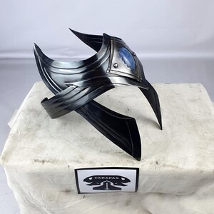 May include: A silver metal headpiece with a blue gemstone in the center. The headpiece has a pointed design and is made of multiple pieces of metal.