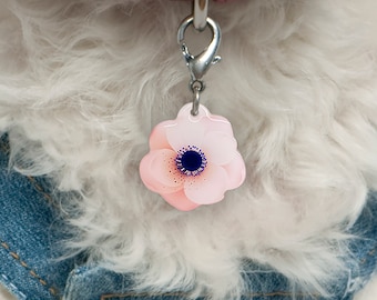 Könnte beinhalten: Ein weißer Hundemarkenanhänger mit einem silbernen Verschluss, verziert mit einer rosa Blume mit blauem Zentrum.