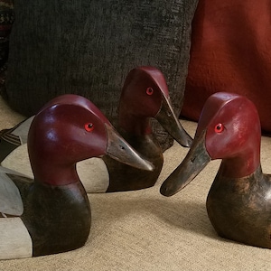 May include: Three wooden duck decoys with red heads, black bodies, and white accents. The decoys are arranged in a semi-circle on a tan surface.