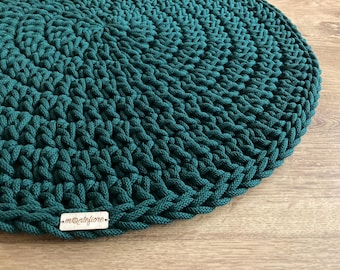 May include: A circular, teal-coloured, crocheted rug. The rug is made of thick yarn and has a textured surface. A small, rectangular tag with the text ".montefiore." is attached to the rug.